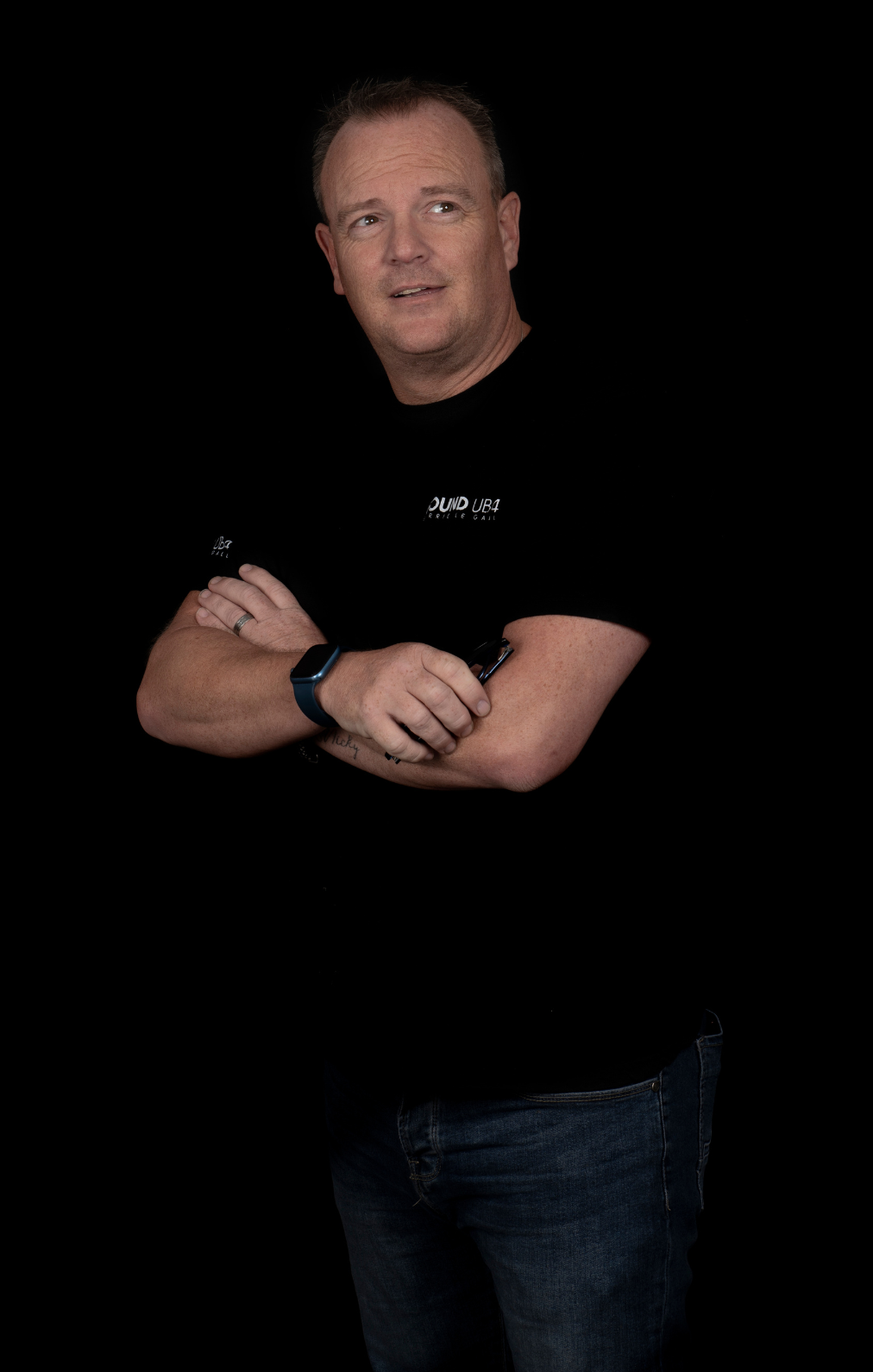 A man with crossed arms wearing a black t-shirt and jeans stands against a black background.