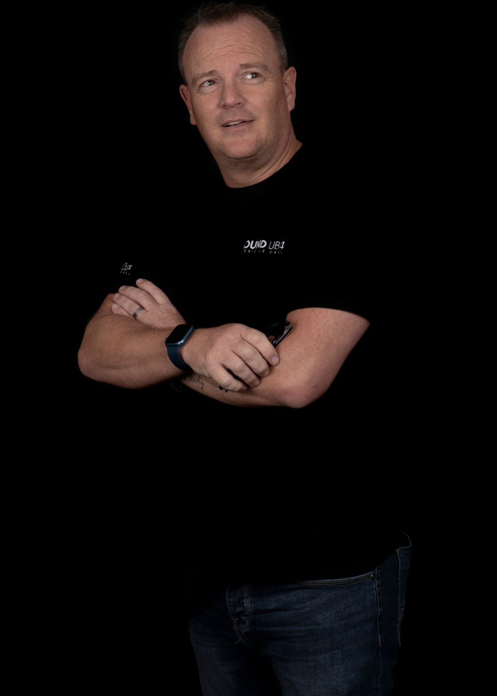 A man with crossed arms wearing a black t-shirt and jeans stands against a black background.